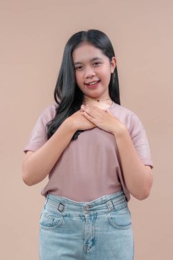 Uzun siyah saçlı, günlük tişört ve kot pantolon giyen Asyalı genç bir kadın. İki eli göğsünde sıcak bir şekilde gülümsüyor. Bej arka planda şükran ve şefkat gösteriyor..