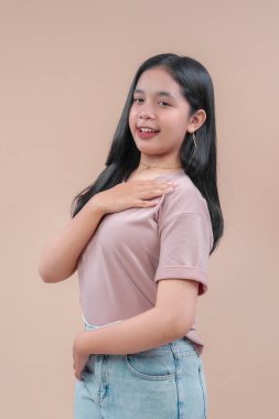 Uzun siyah saçlı, günlük tişört ve kot pantolon giyen, tek eli göğsünde gülümseyen, bej arka planda kendine güvenen ve arkadaş canlısı pozlar veren Asyalı genç bir kadın..
