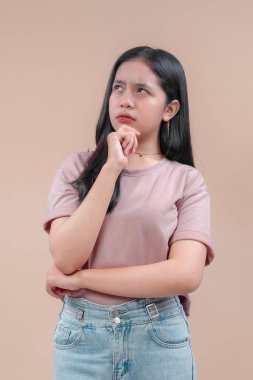 Sıradan modern tarzda, pembe tişört ve kot giyen Asyalı bir kadın, eli çenesinde, meraklı ve odaklanmış bir şekilde düşünceli bir şekilde poz verir..
