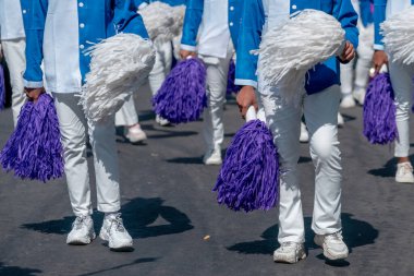 Mavi ve beyaz üniformalı bir grup sanatçı ellerinde mor ve beyaz ponponlar, takım çalışmalarını, kutlamaları ve canlı geçit ruhunu sergileyerek sokaklarda yürüyorlar..