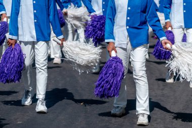 Mavi ve beyaz üniformalı katılımcılar, ellerinde mor ve beyaz ponpon kızlar, takım çalışması, enerji ve şenlikli kutlama ruhunu ifade ederek sokaklarda birlikte yürüyorlar..