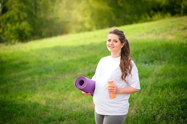 Pregnant woman stands in nature with yoga mat and bottle of water. Pregnant woman before workout