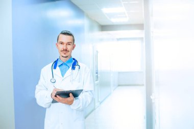 Gözlüklü, kameraya bakan gülümseyen erkek doktor terapisti yakından çek. Hastane koridorunda steteskoplu genç bir doktor.