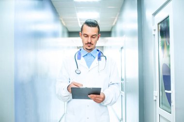 Gözlüklü, kameraya bakan gülümseyen erkek doktor terapisti yakından çek. Hastane koridorunda steteskoplu genç bir doktor.