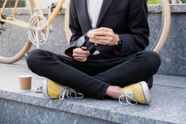 Siyah takım elbise ve rahat ayakkabılar açık havada onun okuma gözlük Temizleme şık adam. Hipster kişi kahve önünde yol bisikleti ile kentsel alanda oturuyor