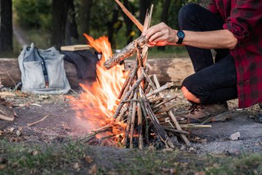 Kamp ateşi bir orman, yapma. Vahşi kavramı içine gidiş: vintage sırt çantası, termos ve erkek rahat kıyafetler içinde kamp yeri ateşe odun parçaları koyar.