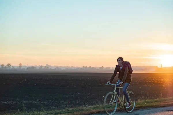 Female commuter riding a bike out of town in rural area. Young woman riding bike at sunset