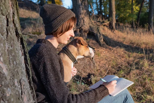 Hiker writing a diary in beautiful nature. Woman facing evening sun takes notes into a notepad, dog sits next to her