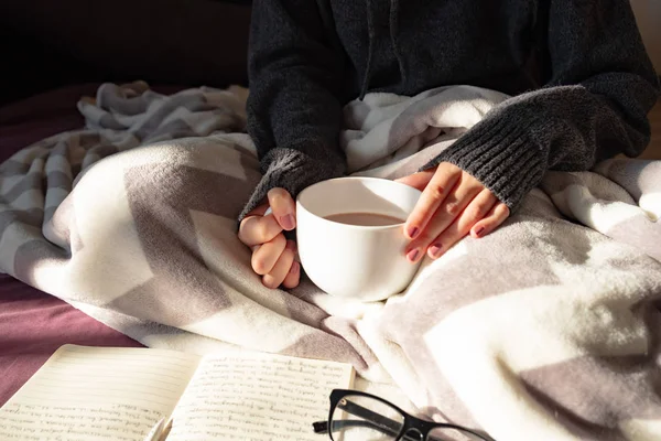 Having coffee and reading in bed on lazy sunny morning. Female hands holding steaming cup of hot cocoa reading in bed