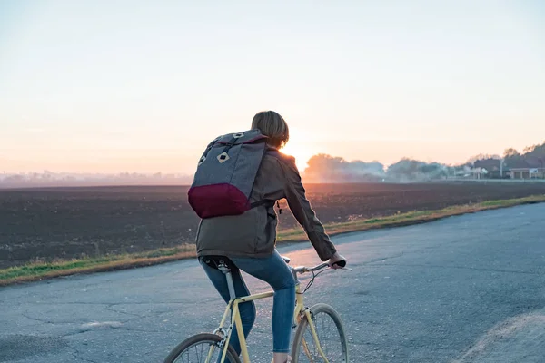 Female commuter riding a bike out of town to a suburban area. Young woman goes home by bike from work along the road at sunset