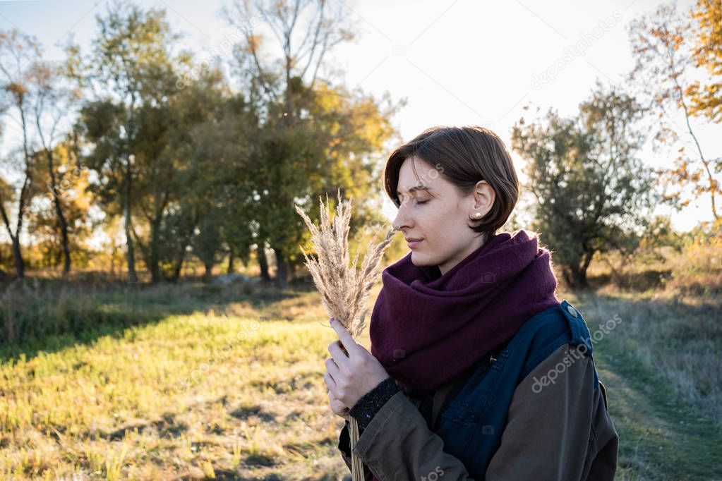 Young woman with a bunch of field grasses enjoying beautiful late summer day. Portrait of female with closed eyes in backlit autumn background