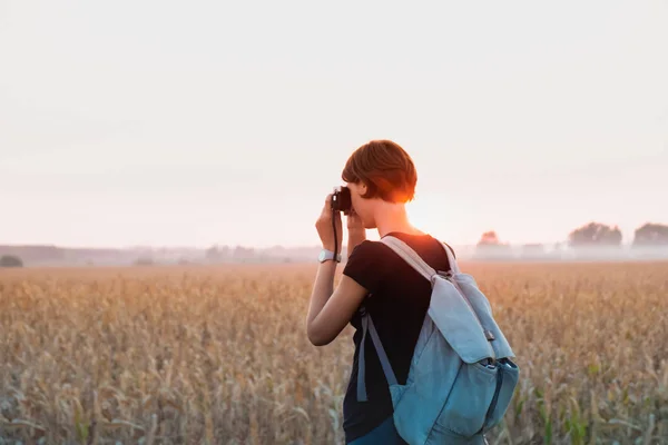 Backlit figure of a woman taking a photo in bright sunset. Female person with backpack standing in evening sunlight at a field and using vintage analog camera