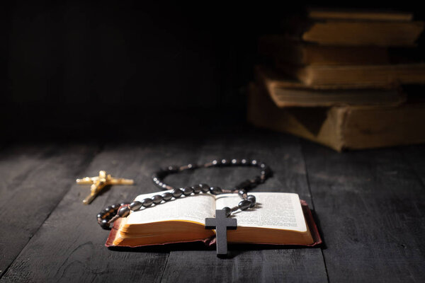 Open book of Bible and crucifix on dark table. Low-key image of New Testament, cross and rosary in bright light among darkness and shadows with copy space