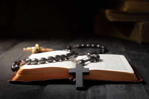 Open book of Bible and crucifix on dark table. Low-key image of New Testament, cross and rosary in bright light among darkness and shadows, close-up view