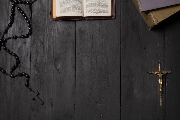 Open book of Bible and crucifix on dark table, top view. Flat lay image of New Testament, cross and rosary on old black background
