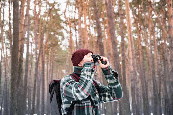 Hiking male person in winter forest taking photograph. Man in checkered winter shirt in beautiful snowy woods uses old film camera