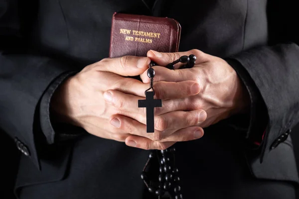 Hands of a christian priest dressed in black holding a crucifix and New Testament book. — Stock Photo, Image