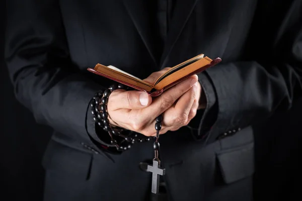 Tangan seorang imam Kristen berpakaian hitam memegang salib dan membaca buku Perjanjian Baru . — Stok Foto