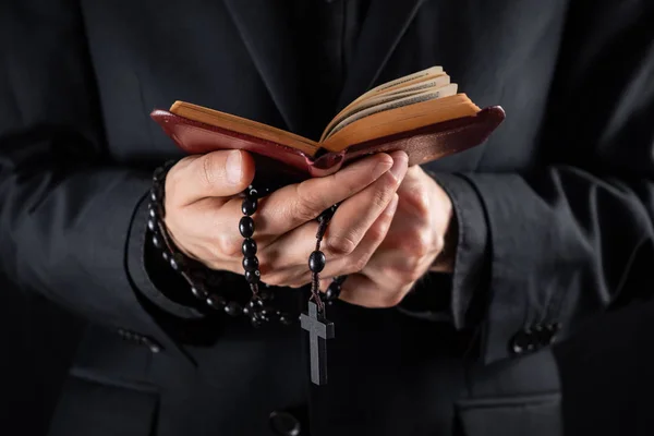 Hands of a christian priest dressed in black holding a crucifix and reading New Testament book — Stock Photo, Image