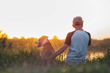 Adam gün batımında göl kenarında köpeğine sarılıyor. Evcil hayvanlar ve insan dostluğu, ilgilenmek, birlikte zaman geçirmek..