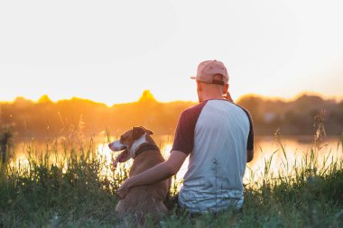 Adam gün batımında göl kenarında köpeğine sarılıyor. Evcil hayvanlar ve insan dostluğu, ilgilenmek, birlikte zaman geçirmek..