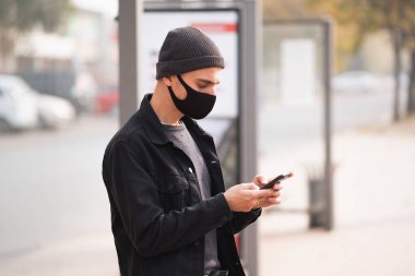 Otobüs durağında cep telefonu olan maskeli bir adam. Millennial halka açık bir yerde akıllı telefon kullanıyor.