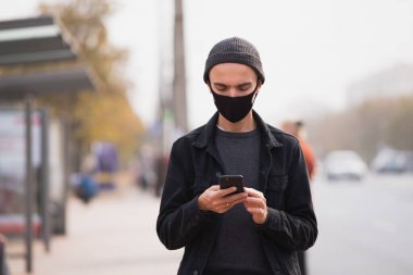 Otobüs durağında cep telefonu olan maskeli bir adam. Millennial halka açık bir yerde akıllı telefon kullanıyor.