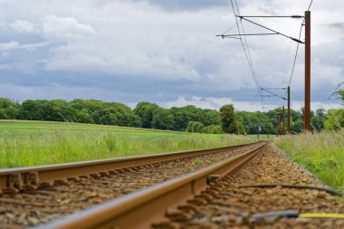 Paslı metal raylar taştan bir yatakta ilerler. Elektrik kablolarını tutan sırıklar. Tarlalar ve çimenler her iki tarafa da yayıldı ve pist perspektifi görüntüyü mesafeye götürüyor..