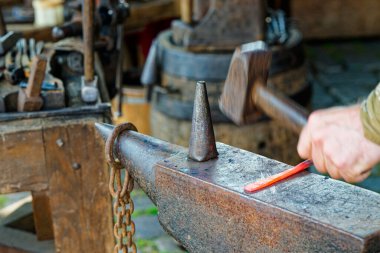 Demirci metali ısıtırken zincirler bankın arkasında asılı duruyor. Aletler istasyon hizasında, ve stoktaki turuncu parıltı değişim, güç ve ateşin ustalıkla birlikte çalıştığını gösteriyor..
