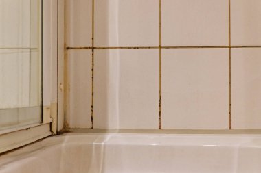 Parlak beyaz bir duş odası perde yerine sürgülü cam bir ekran kullanır. Döşeme eklemleri ağır siyah küf ve nem lekeleri gösteriyor. Temiz düzen, kötü havalandırmayı hatırlatan sert bir şeye dönüşüyor..