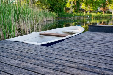 Dar bir ahşap iskele durgun suyun üzerine uzanır, küçük plastik bir tekne kalasların yanında demirler, göl cam gibi sakindir, kıyı yakındır ve yansımaları naziktir, korunaklı bir gölette sakin bir duraksama.