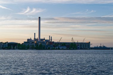 Deniz kıyısındaki Stadtwerke Flensburg kompleksinden limana bakan uzun vinçler rıhtımda ve yakınlarda demirlemiş yatlarla. Metal çerçeveler, limanlar ve direkler etkin bir sanayi kıyı şeridi çiziyor.