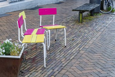 Bir saksının yanındaki kaldırımda sarı ve pembe tonlu üç küçük boyalı sandalye bir bankın yanında duruyor ve neşeli bir şehir manzarası yaratıyor..