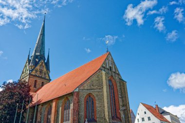 Keskin kulesi olan uzun gotik bir kiliseye açılan bir Alman kasabası. Küçük kanat tepeleri yüksekliği yansıtıyor. Yanında yuvarlak pencereli bir kilise kanadı var. Açık mavi gökyüzü görüntüyü tamamlar.