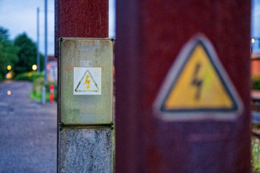 İki aşınmış direk bir sınırda duruyor. Birinde yüksek voltaj işareti var, diğerinde de aynı sembolle gri bir servis kutusu var. Paslı pullar ve kirli lekeli hava yılları.