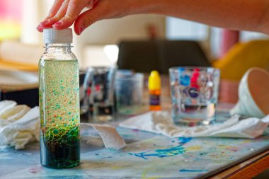 Pembe tırnaklı bir kız açık bir şişe tutuyor, sarı başlıklı ve beyaz kapaklı, kırmızı ve mavi renkli kabarcıklar içinde, derin yeşil bir katman ise eğlenceli bir ev bilimi anı için aşağıda duruyor..