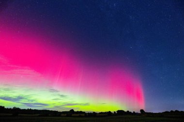 Yoğun bir aurora geceyi kaplıyor. Uzun perdeler sessiz bir tarlayı süpürüyor ve gökyüzünü parlak bir renk sarıyor. Açık kırsal bölgede sayısız yıldız parlıyor..
