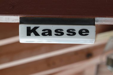Basit ahşap bir duvar üzerinde Kasse yazan küçük bir tabela taşır. Açık bir mekanda nakit masası ya da bilet noktasını işaret eder. Sıcak dokular festival pazarları ve etkinlik markalaşmasına uygun.