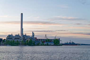 Suyun karşısında Rheinmetall kompleksi limana bakıyor. Rıhtımdaki ağaçlar ve iskelenin yanındaki demirli yatlar. Yakınlarda bir sanayi gemisi duruyor. Yoğun ama sakin bir kıyı sahnesini tamamlıyor..