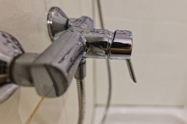 Beyaz fayanslı duş duvarları, su damlacıkları tarafından benekli esnek bir hortumla krom bir musluk çerçevesi oluşturuyor. Hijyen temalarına, kaplıcalara, ürün modellerine ve evde bakım reklamlarına uyan temiz bir tuvalet ayrıntısı..