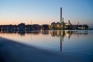 Liman lambaları sakin yataklar üzerinde titreşir. Rheinmetall yığını yeşil parlarken, dükkan önleri ve vinçler körfezde şerit olarak yankılanıyor. Yatlar sabit durur, ince inşaat perdeleri alacakaranlıkta kaybolur..