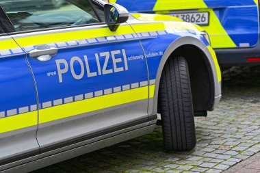 Schleswig Holstein 'in kuzeyinde iki gri arazi aracı mavi ve sarı işaretler gösteriyor. En yakın araç önde duruyor, diğeri ileride bekliyor, çift resmi ve temiz bir kaldırımda görünüyor..