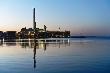 Akşam, metal fabrikasının yanındaki limana yerleşir, lambalar parlar ve bir yığın yeşil parlar. Işıkları durgun sulara yayılıyor mavi ve gül renginde, endüstriyi sessiz bir sahneye dönüştürüyor..