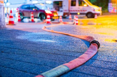 Sırılsıklam olmuş asfaltta bir yangın hortumu düğümleniyor. Ötede iki kırmızı araba ve bir kurtarma aracı bekliyor ve beyaz ve kırmızı bir sıra şeridi işaret ediyor. Yolda ve hortumda boncuklar parıldıyor..