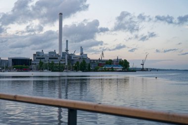 Gün ışığı limandaki renkleri düzleştirir. Gri bulutlar alçaktan sarkır, su küçük dalgaları kaldırır ve rıhtımda yatlar kümelenir. Stadtwerke Flensburg 'un çalışmaları ışıksız ve sade bir şekilde yükselir..