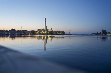 Akşam Flensburg limanında. Demirlemiş yatlar düzenli sıralarda dinleniyor ve Rheinmetall bacası yeşil parlıyor, suyun üzerinde ışık dalgaları oluşuyor. Uzak alan ekran hattı uzaktaki rıhtımda.