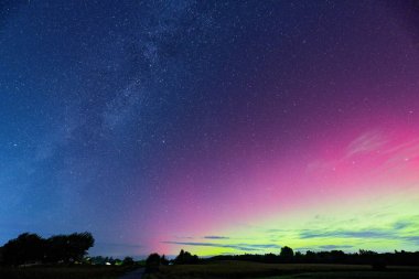 Açık bir alan yıldızlardan oluşan bir duvar halısının altında uzanırken, kuzey ışıklarının geniş yayları ufuk boyunca hafif renk değişimleriyle gökyüzünü kaplıyor ve berrak gece manzarası huzurlu hissettiriyor..