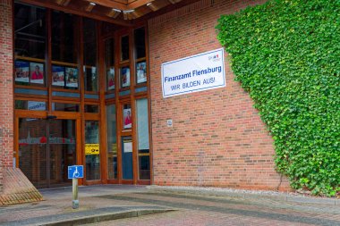 Kırmızı cephede Finanzamt Flensburg yazan beyaz bir plaket var. Tekerlekli sandalye sembolü ziyaretçiler için girişi gösteriyor. Giriş resmi ve düzenli görünüyor, etrafında basit süslemeler ve düzgün tuğlalar var..