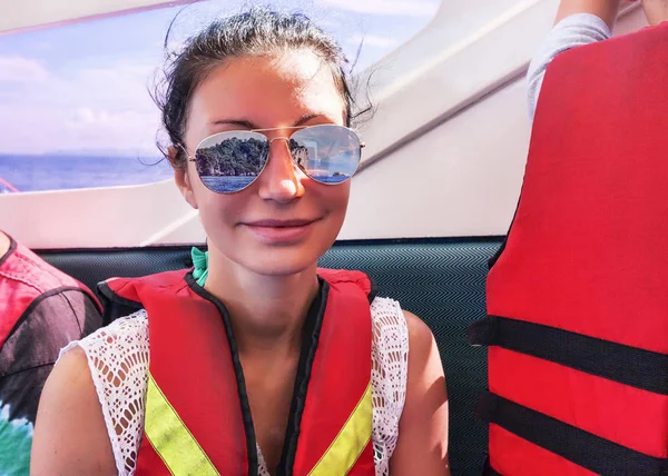 Smiling dark-haired woman sails on tropical sea wearing lifejacket and sunglasses