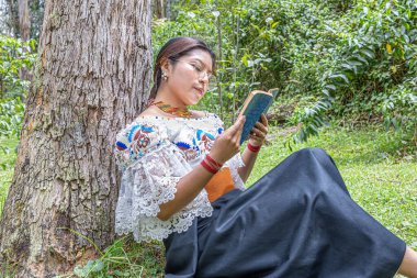 Otavalo, Ekvador: 6 Ağustos 2025: Geleneksel işlemeli giysiler içinde çıplak ayaklı yerli Otavalo kadını, doğal bir And ortamında bir ağaç gövdesine yaslanırken bir kitap okuyor, barış ve kültürel bağlantı kuruyor..
